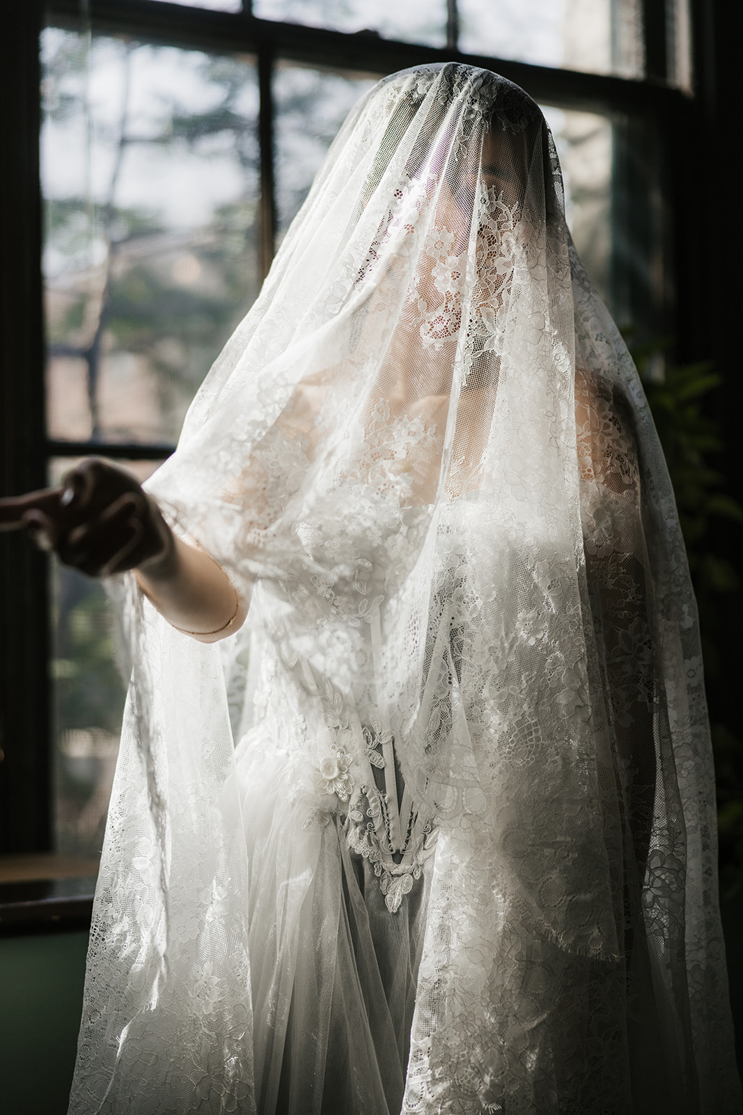A person wearing a wedding dress and veil stands in front of large windows, illuminated by natural light.