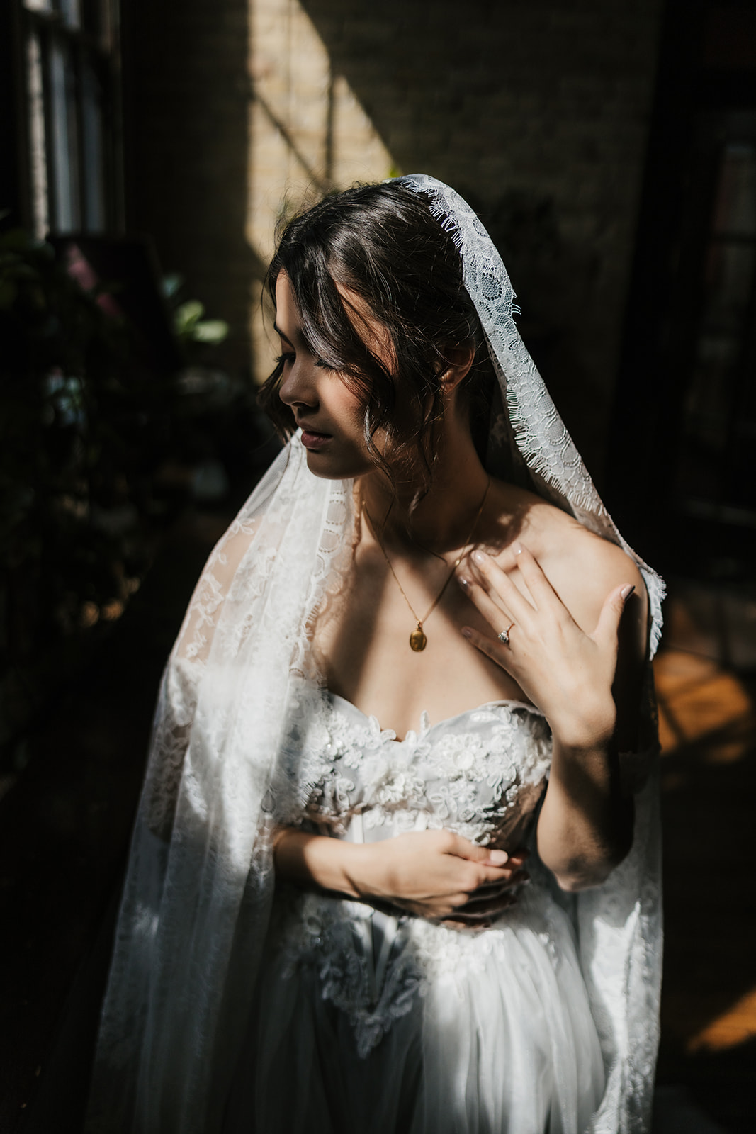 A person wearing a wedding dress and veil stands in front of large windows, illuminated by natural light.