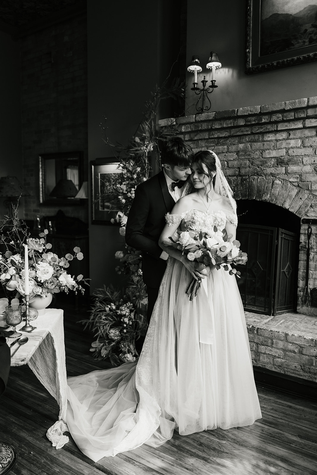 A bride in a white dress holding a bouquet stands next to a groom in a black suit in front of a brick wall decorated with flowers.