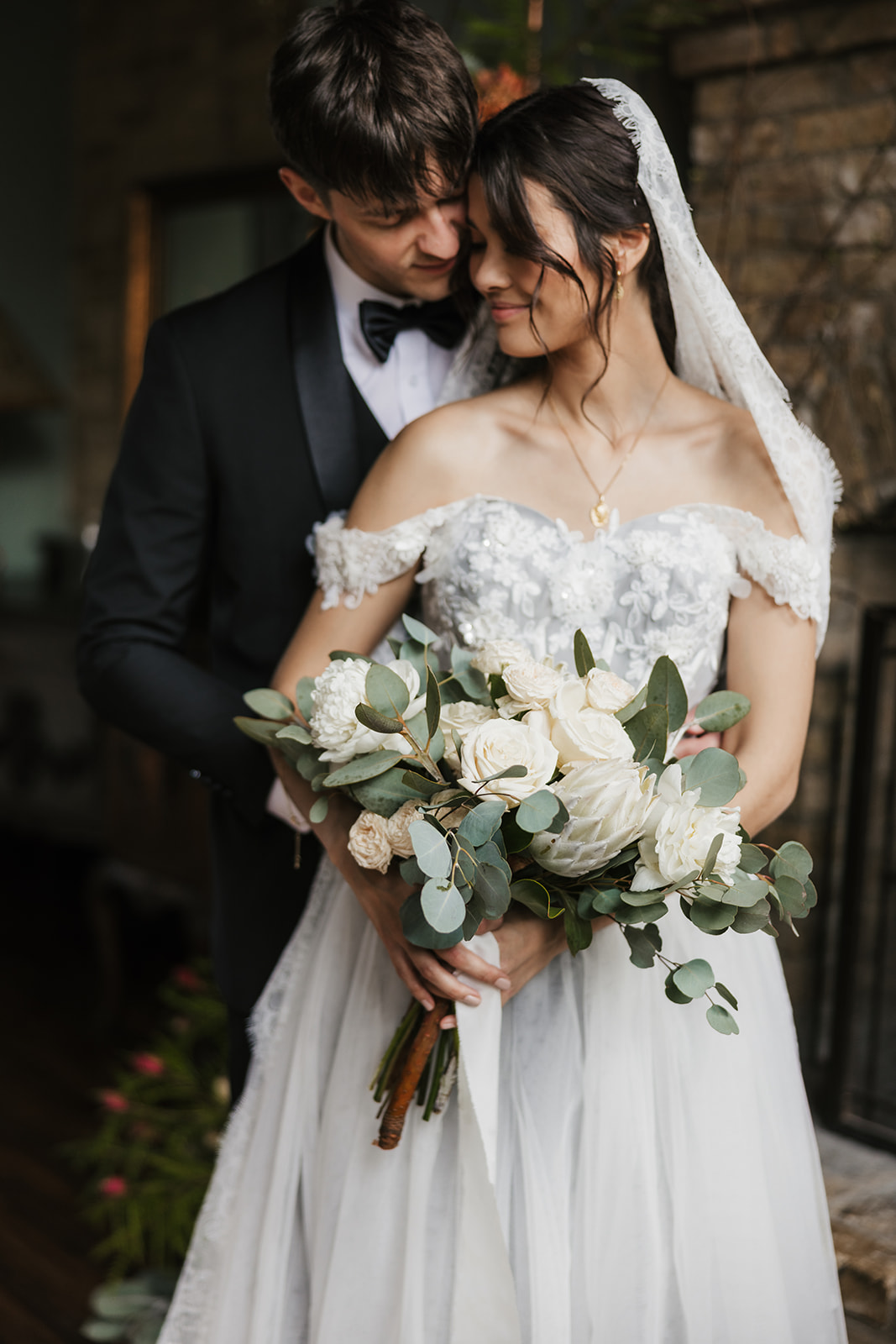 A bride in a white dress holding a bouquet stands next to a groom in a black suit in front of a brick wall decorated with flowers.