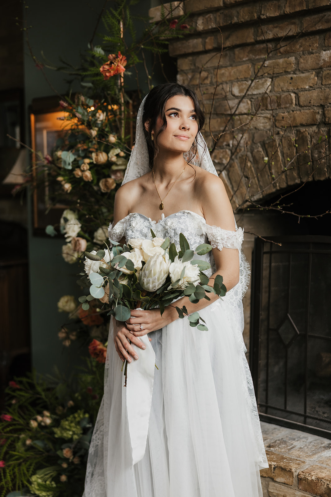 A bride in a white dress holding a bouquet stands next to a groom in a black suit in front of a brick wall decorated with flowers.