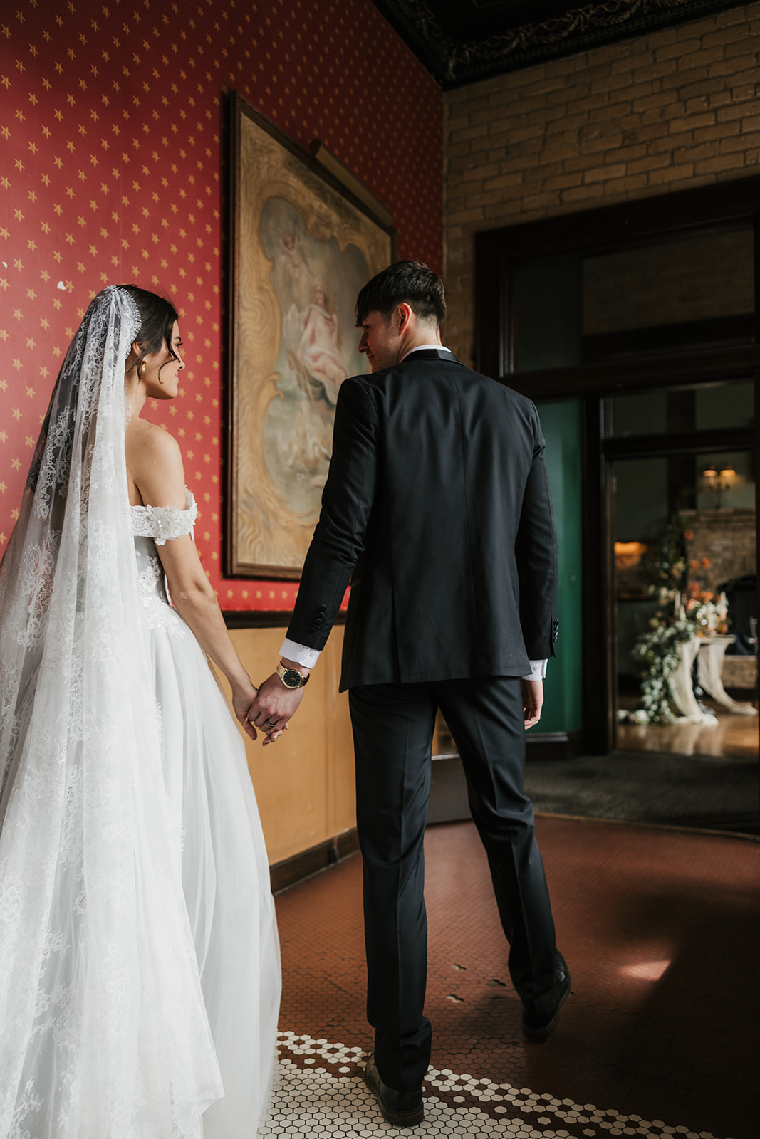 A bride and groom in formal attire hold hands and walk away together in an elegant indoor setting.