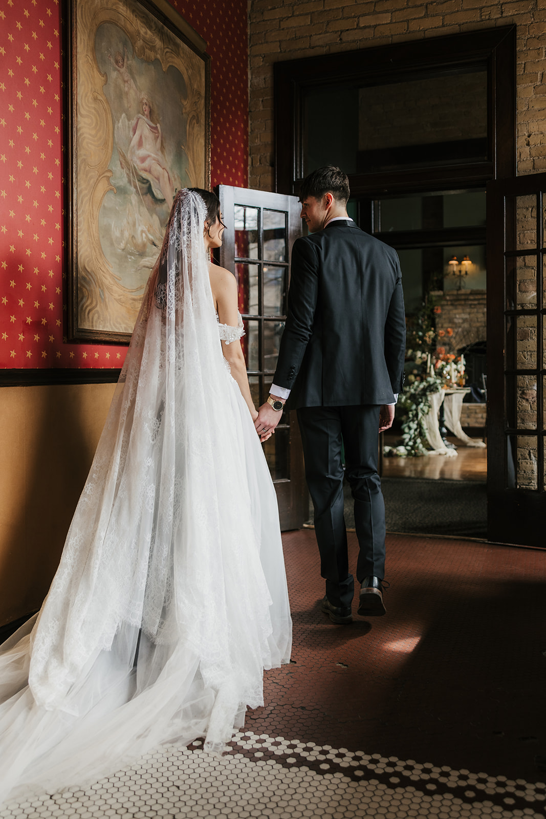 A bride and groom in formal attire hold hands and walk away together in an elegant indoor setting.