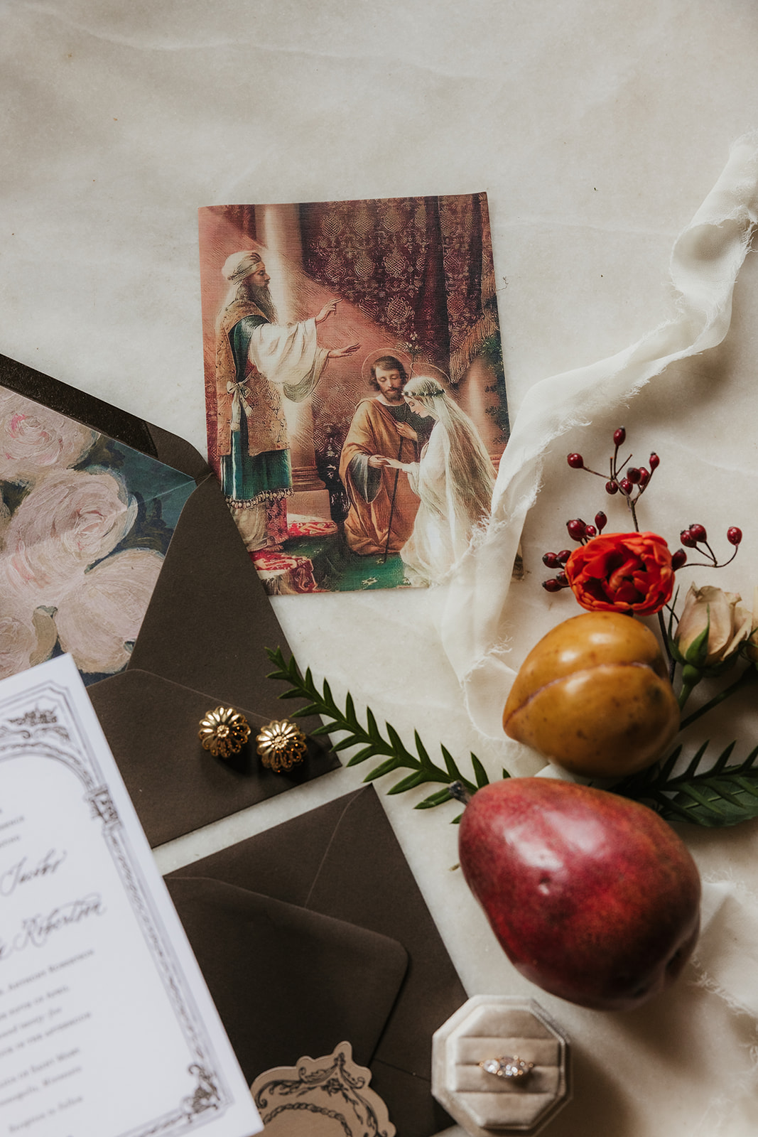 A flat lay of vintage wedding stationery with invitations, envelopes, a rosary, pearls, grapes, wax seal, and decorative flowers arranged on a marble surface for a catholic wedding idea