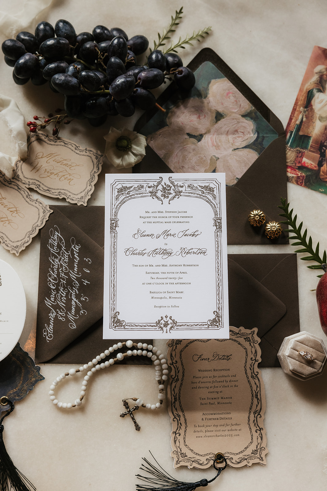 A flat lay of vintage wedding stationery with invitations, envelopes, a rosary, pearls, grapes, wax seal, and decorative flowers arranged on a marble surface for a catholic wedding idea