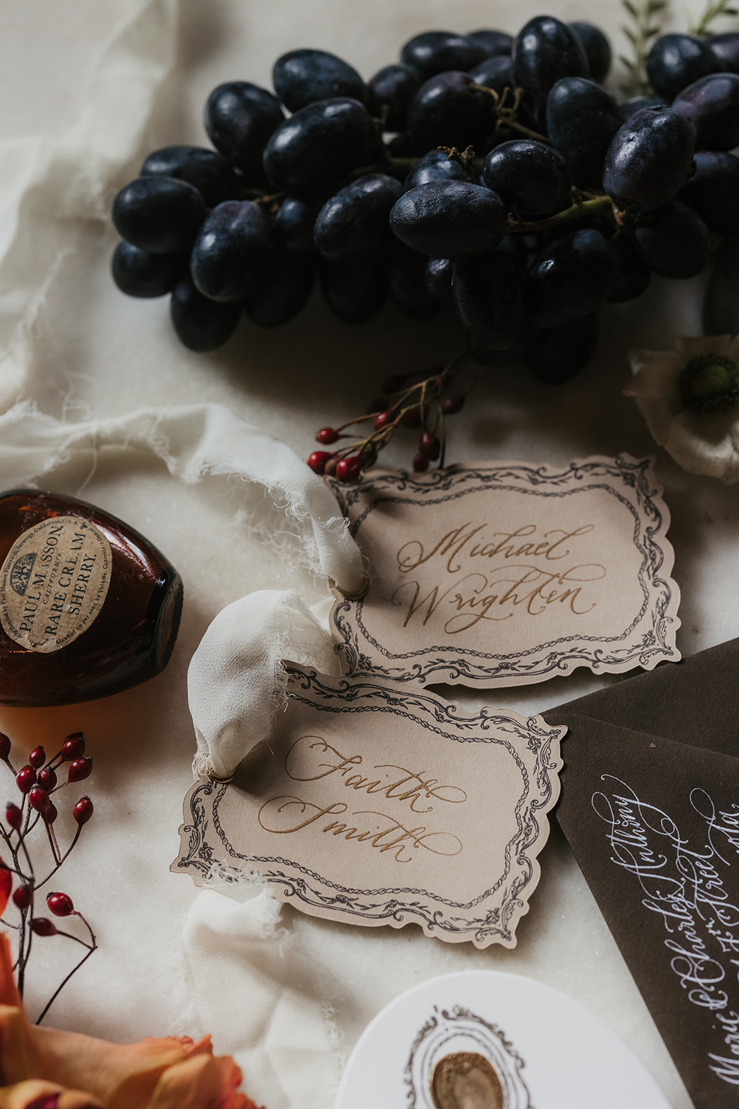 A flat lay of vintage wedding stationery with invitations, envelopes, a rosary, pearls, grapes, wax seal, and decorative flowers arranged on a marble surface for a catholic wedding idea