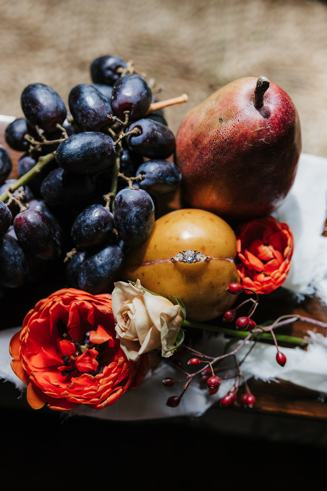 A still life arrangement featuring dark grapes, a pear, yellow fruits, orange and white flowers, and red berries on a white cloth.