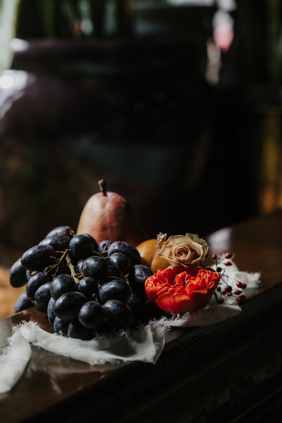 A still life arrangement featuring dark grapes, a pear, yellow fruits, orange and white flowers, and red berries on a white cloth.