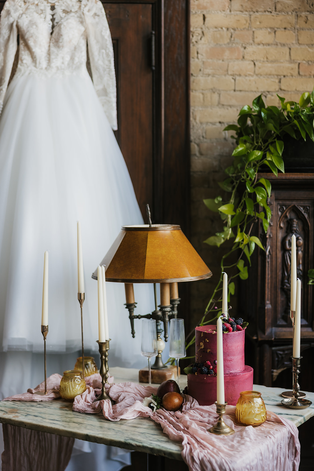 A long-sleeved wedding dress hangs on a door next to a table set with candles and decor in a room with brick walls.
