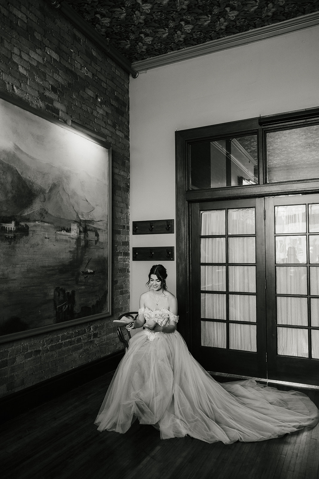 A woman in an off-the-shoulder white wedding dress sits on a chair and holds a pair of beige high heels in her hand for catholic wedding ideas