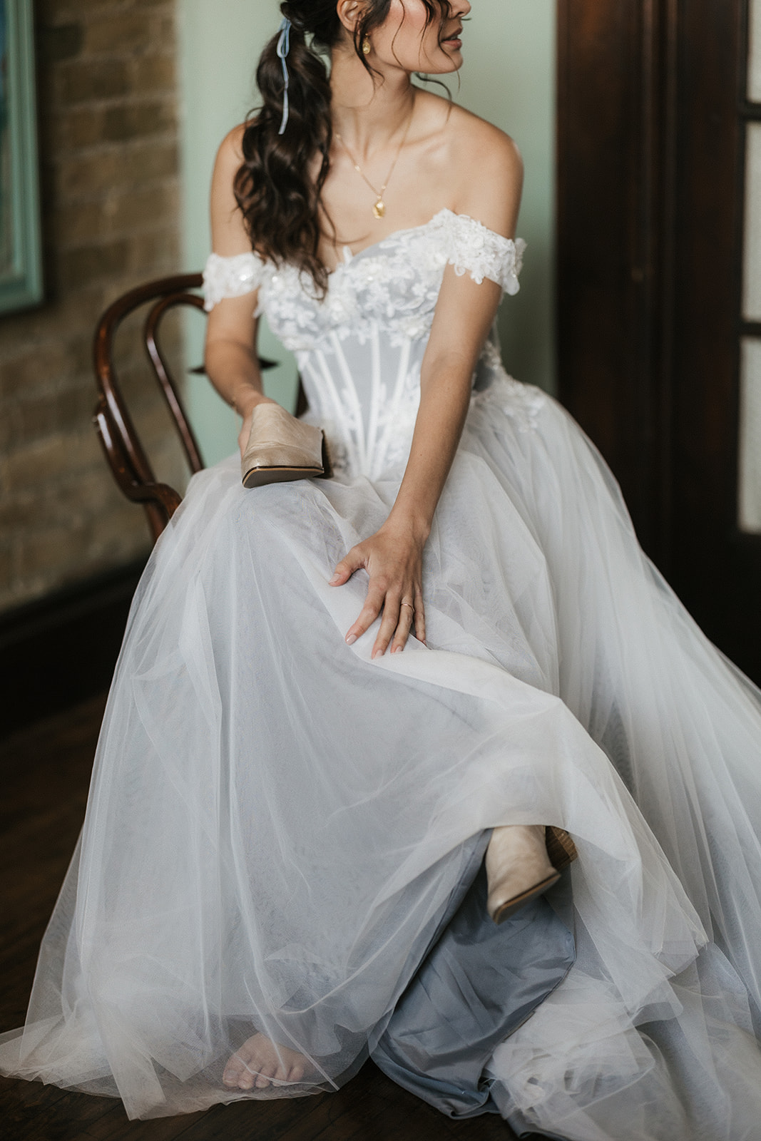 A woman in an off-the-shoulder white wedding dress sits on a chair and holds a pair of beige high heels in her hand for catholic wedding ideas