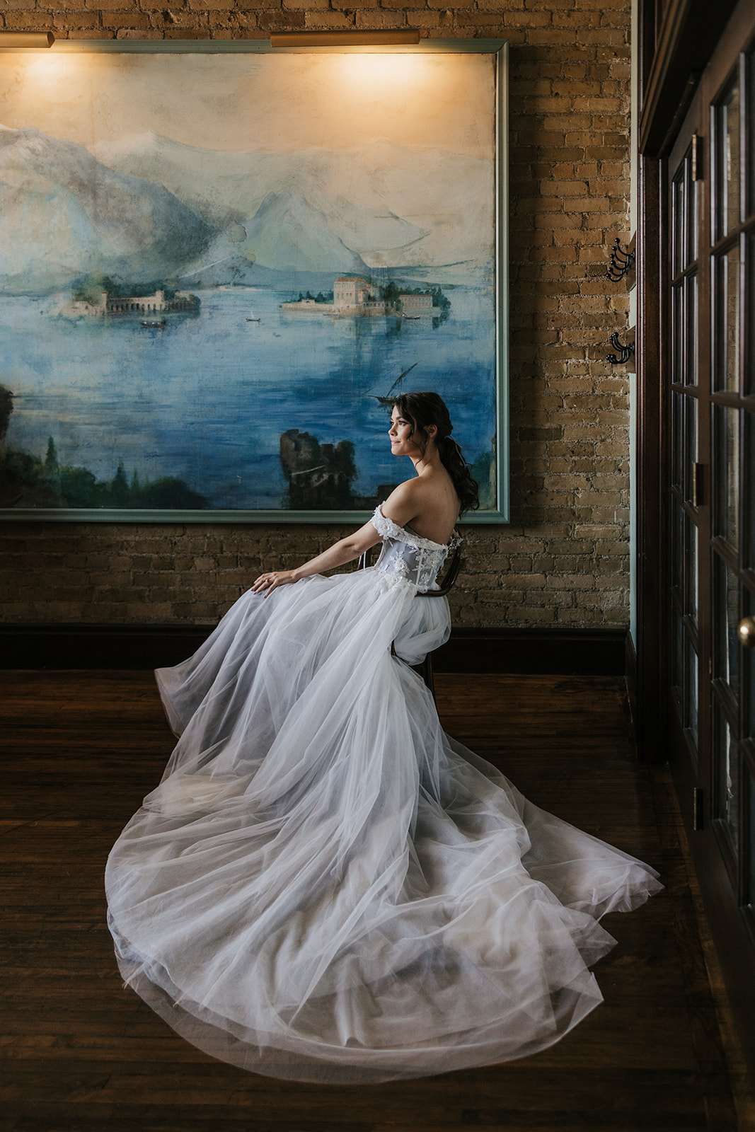 A woman in an off-the-shoulder white wedding dress sits on a chair and holds a pair of beige high heels in her hand for catholic wedding ideas