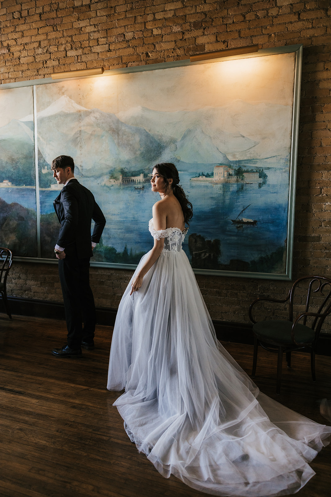 A bride in a white gown and a groom in a black suit stand indoors in front of a large painting on a brick wall.