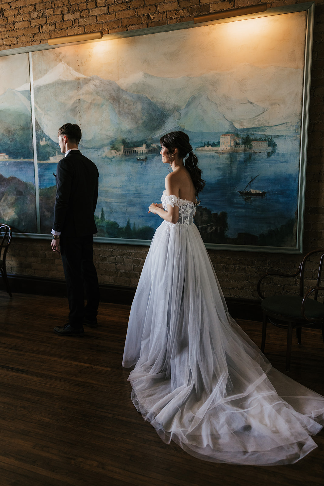 A bride in a white gown and a groom in a black suit stand indoors in front of a large painting on a brick wall.