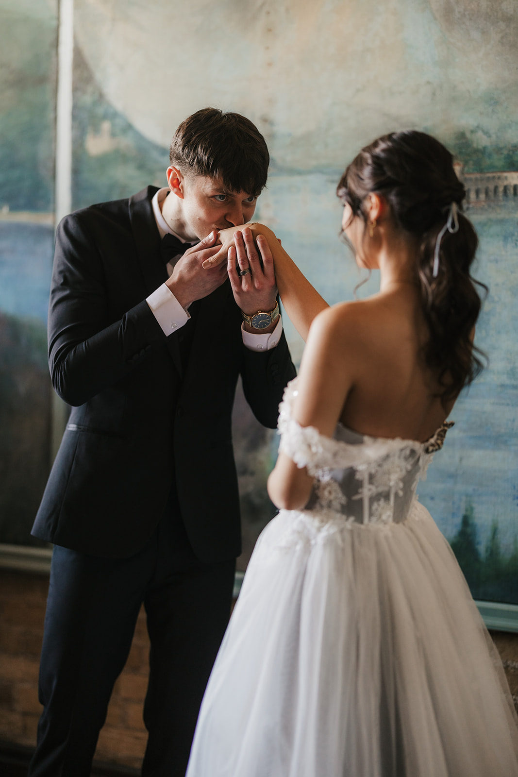 A bride in a white gown stands beside a seated groom in a dark suit, with a painted mural of mountains and water in the background for catholic wedding ideas