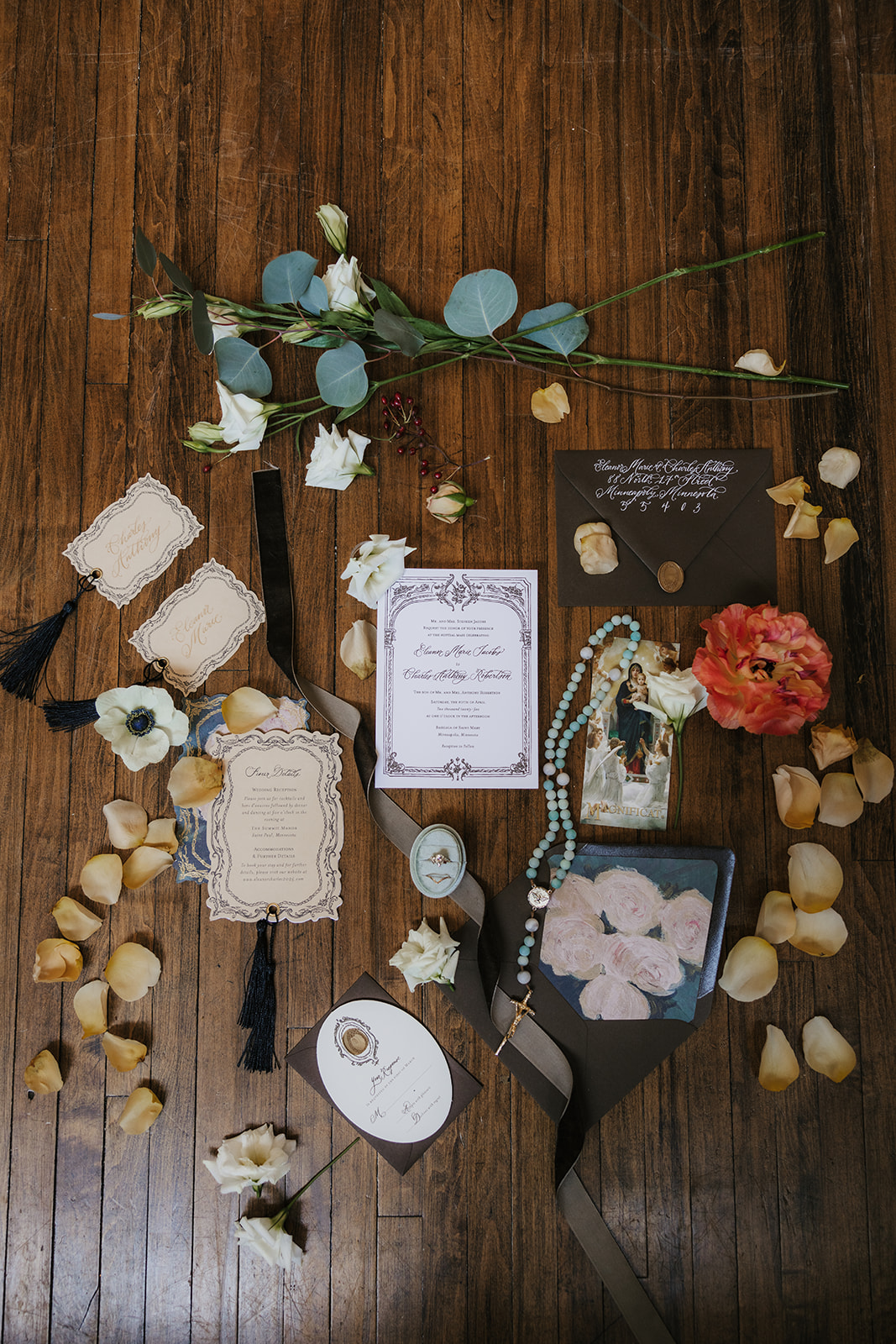 Flat lay of wedding invitations, floral arrangements, a beaded necklace, a photo, and decorative items arranged on a wooden floor for catholic wedding ideas