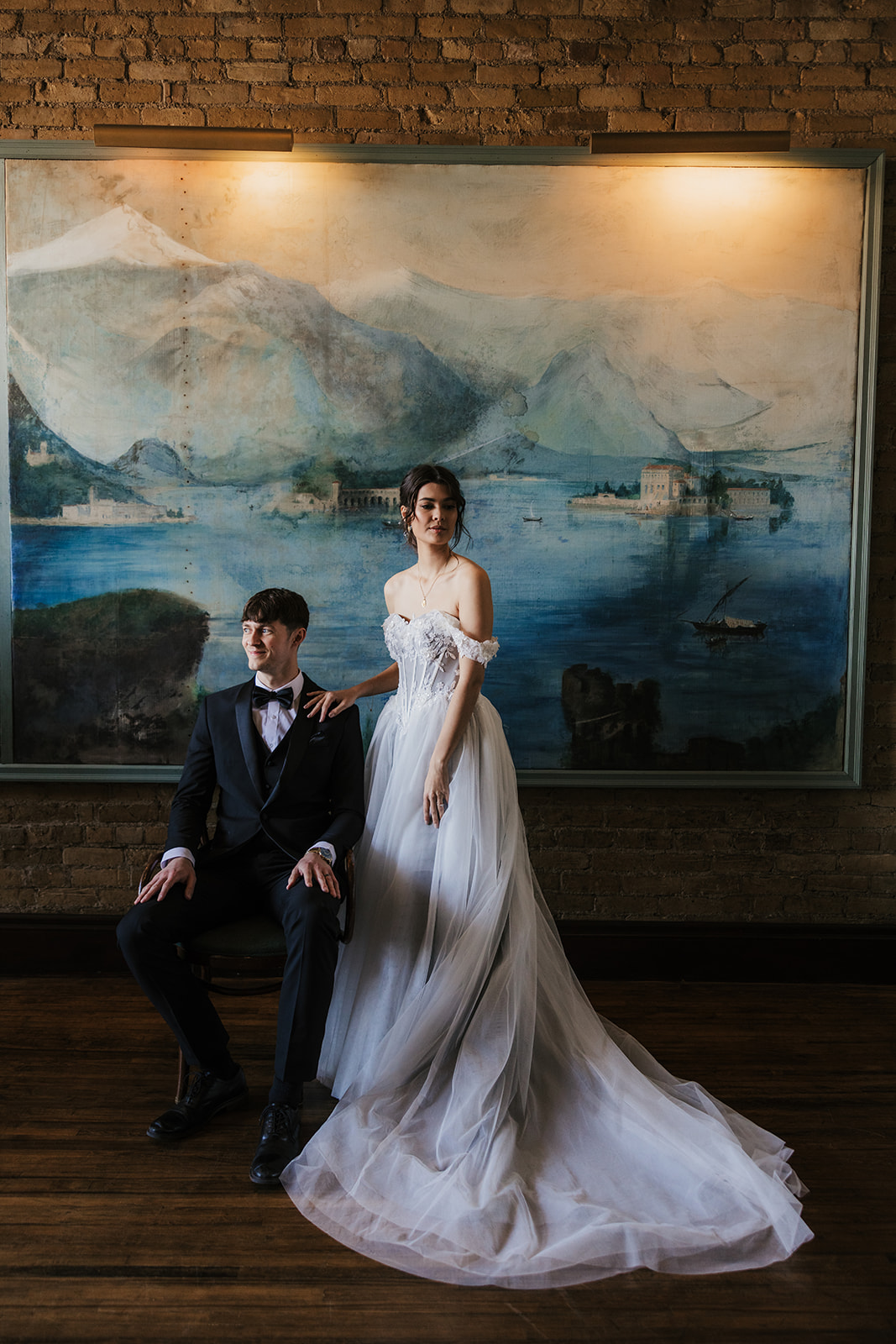 A bride in a white gown stands beside a seated groom in a dark suit, with a painted mural of mountains and water in the background for catholic wedding ideas