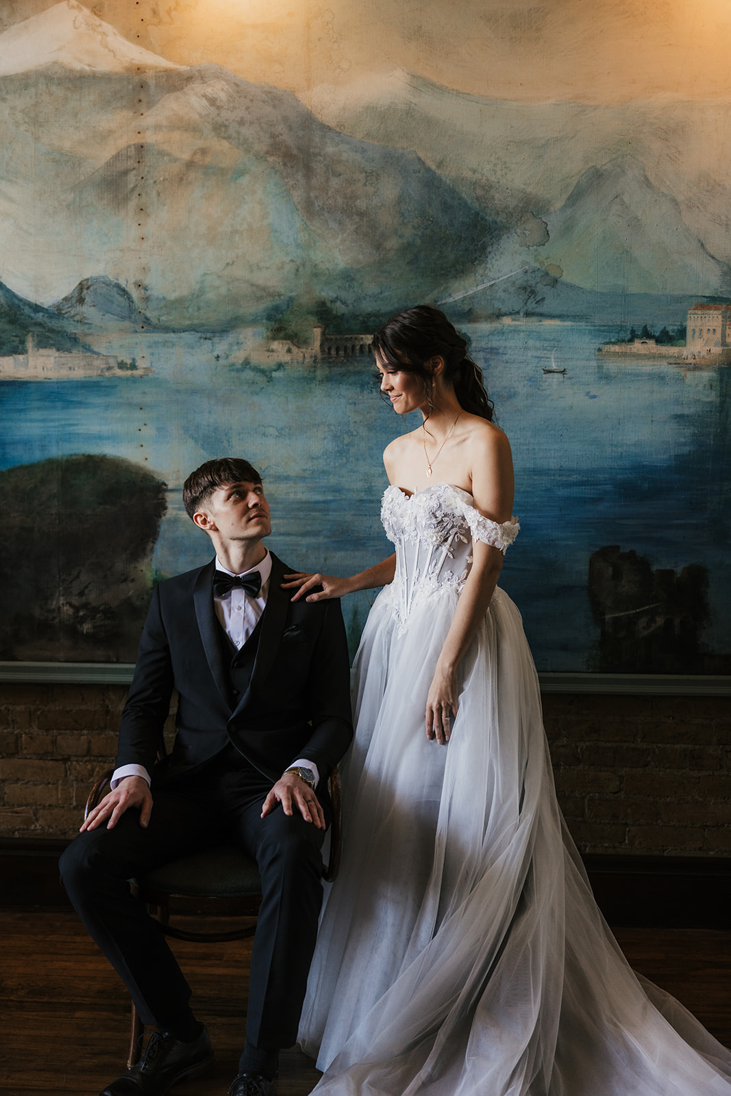 A bride in a white gown stands beside a seated groom in a dark suit, with a painted mural of mountains and water in the background for catholic wedding ideas