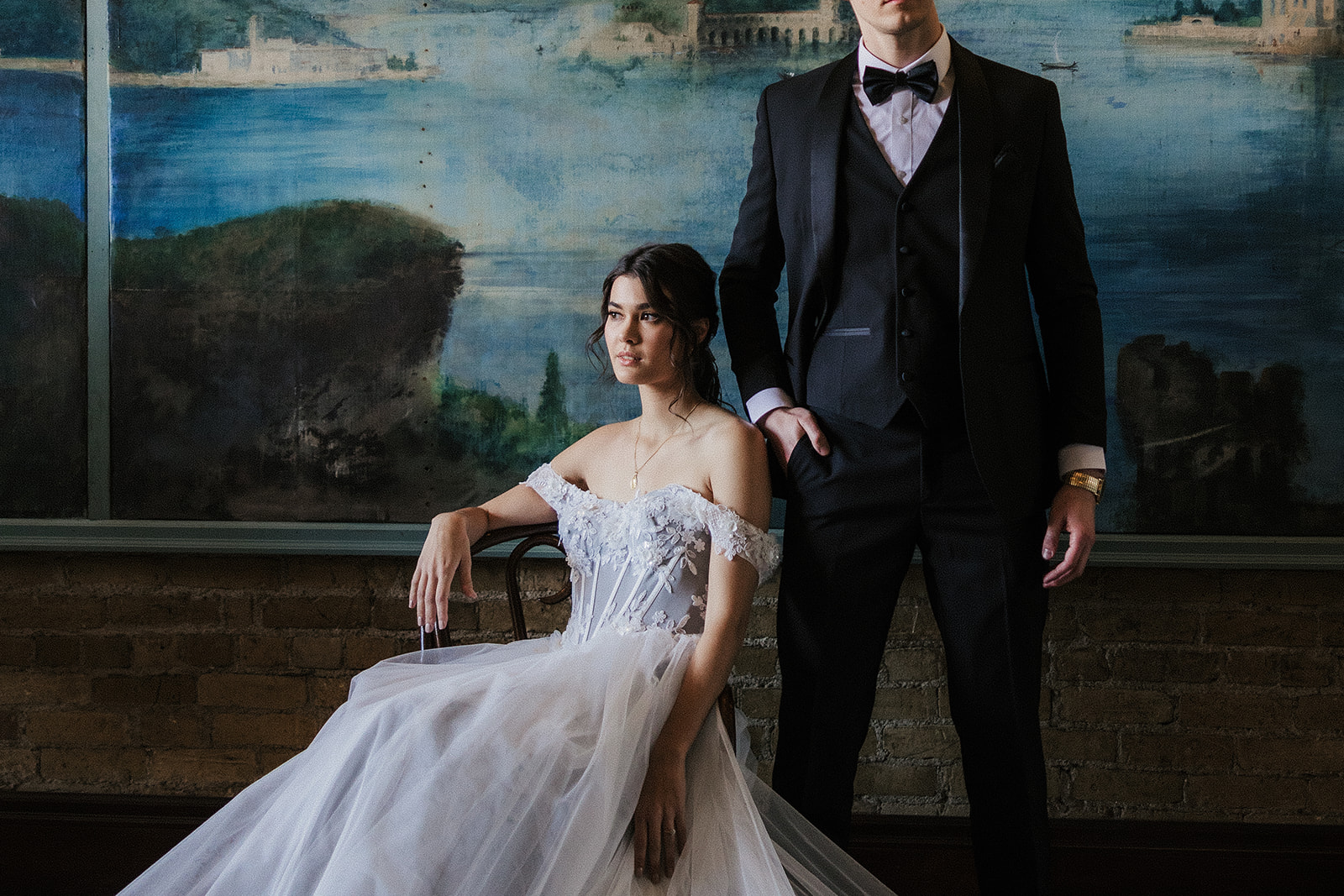 A bride in a white gown stands beside a seated groom in a dark suit, with a painted mural of mountains and water in the background for catholic wedding ideas