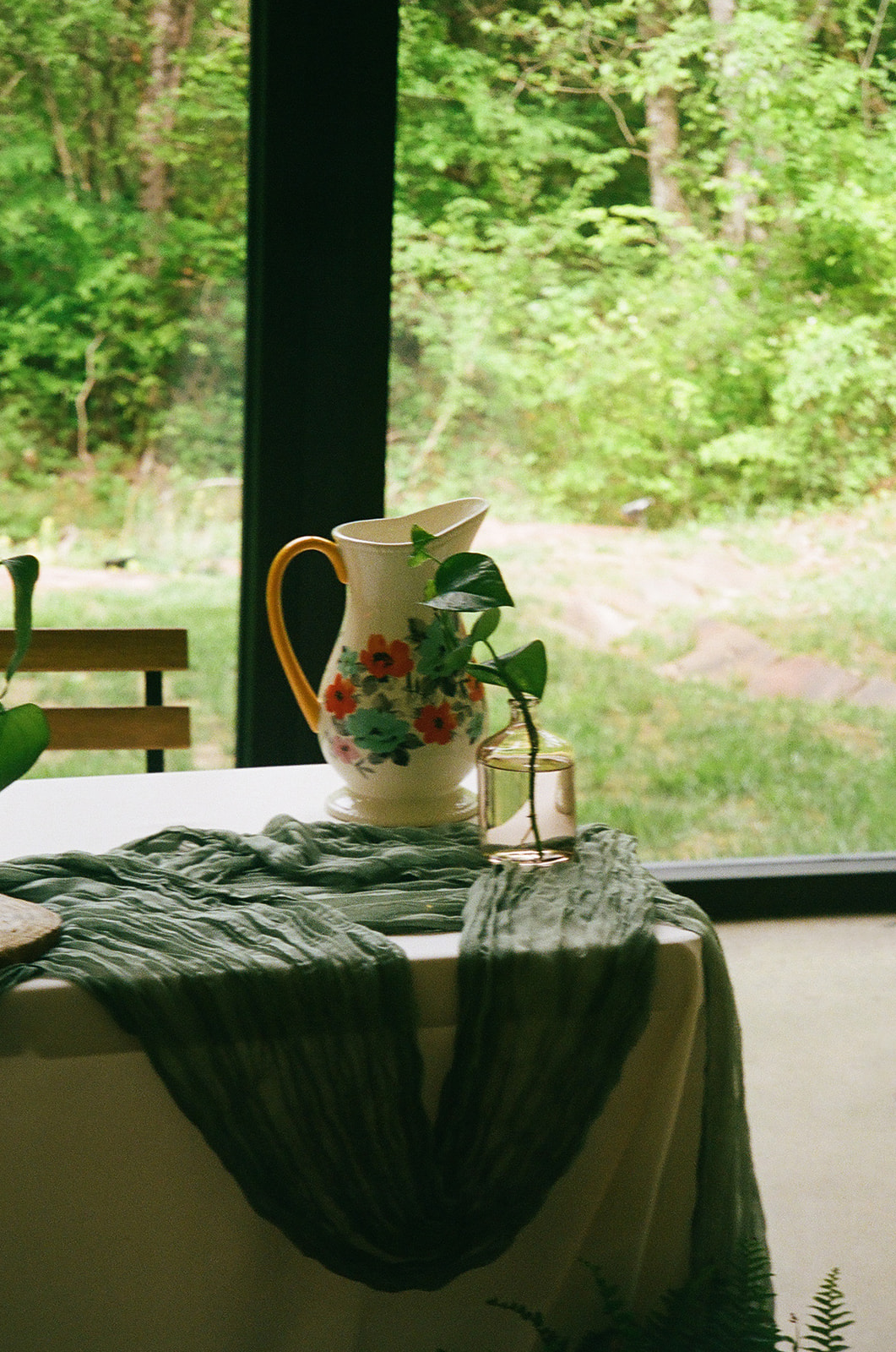 Rectangular tables with white tablecloths and green runners are arranged in a bright room with large windows and wooden chairs, overlooking a green outdoor view.