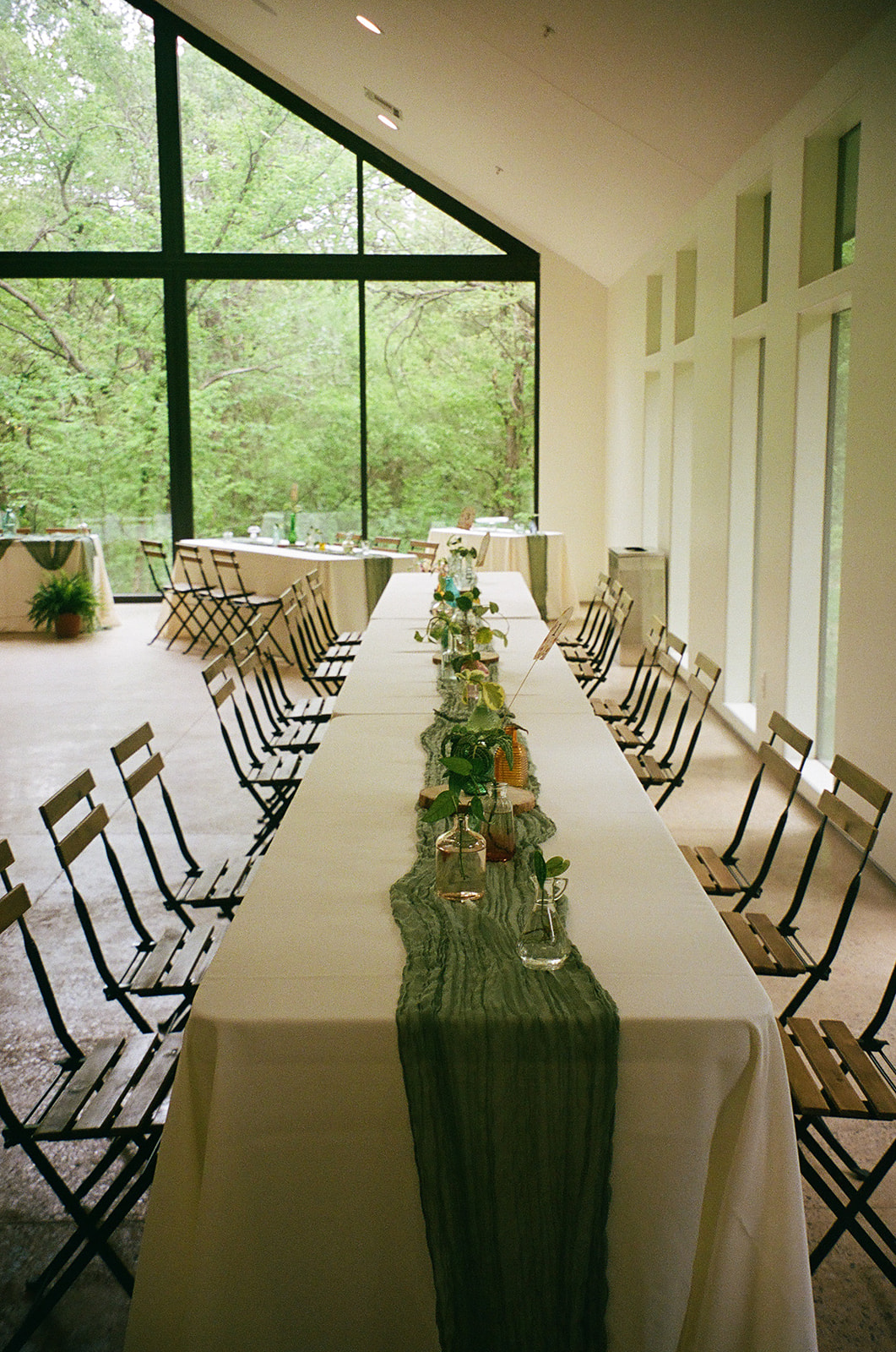 Rectangular tables with white tablecloths and green runners are arranged in a bright room with large windows and wooden chairs, overlooking a green outdoor view.