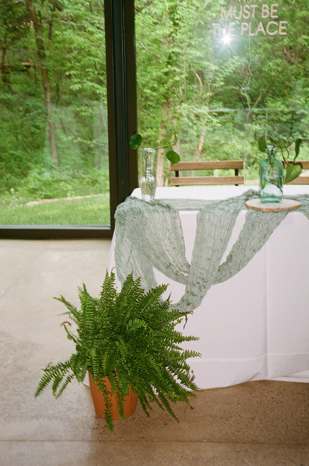 Rectangular tables with white tablecloths and green runners are arranged in a bright room with large windows and wooden chairs, overlooking a green outdoor view.