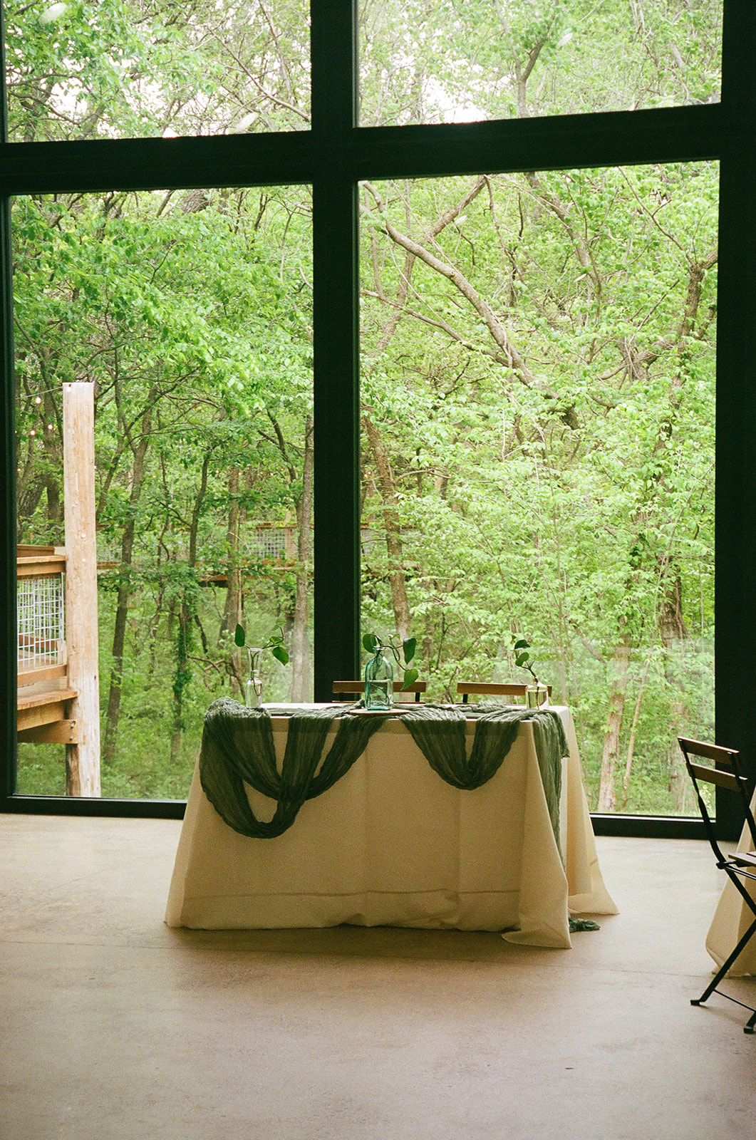Rectangular tables with white tablecloths and green runners are arranged in a bright room with large windows and wooden chairs, overlooking a green outdoor view.