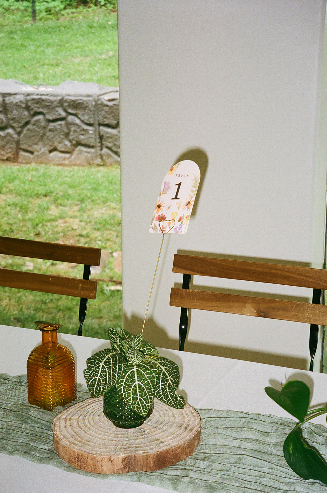 Rectangular tables with white tablecloths and green runners are arranged in a bright room with large windows and wooden chairs, overlooking a green outdoor view.