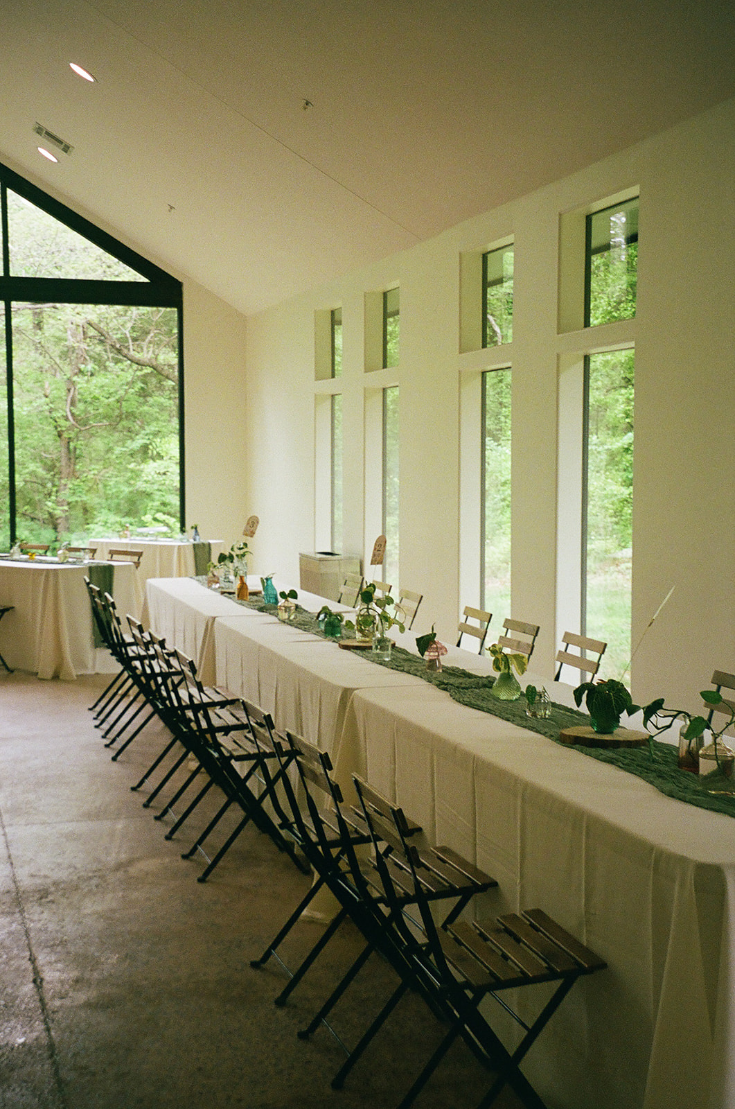 Rectangular tables with white tablecloths and green runners are arranged in a bright room with large windows and wooden chairs, overlooking a green outdoor view.