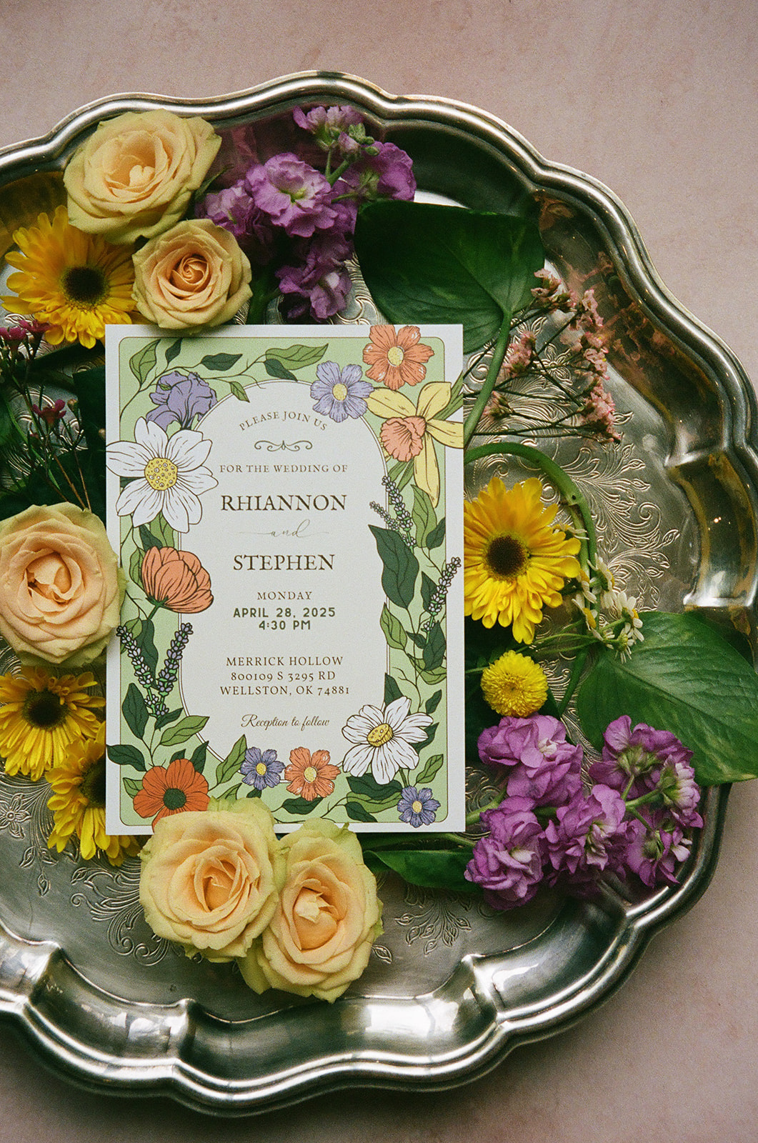 Two floral wedding invitations surrounded by yellow and purple flowers, green leaves, a wax seal, and a decorative box arranged on a light pink surface.