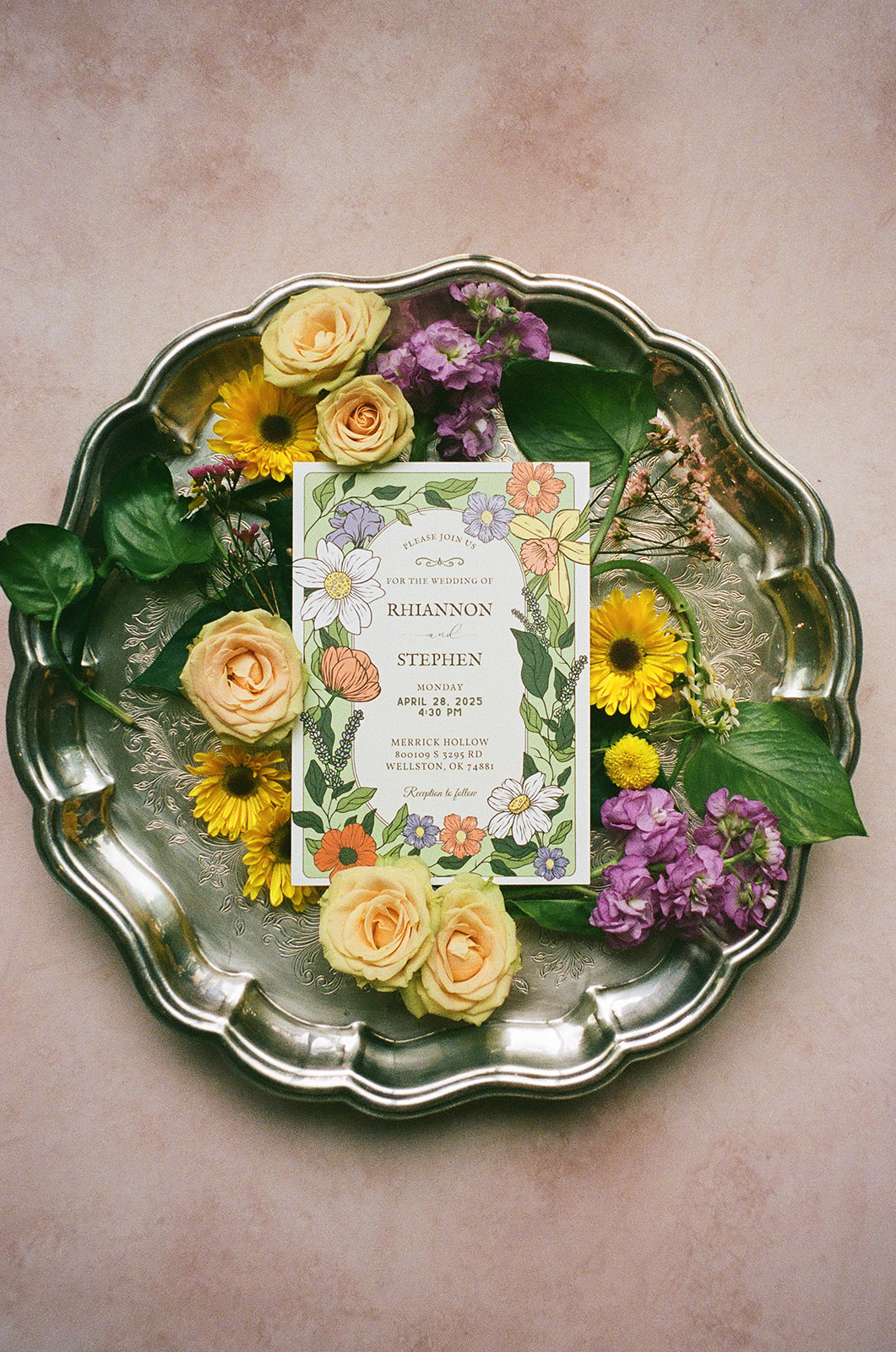 Two floral wedding invitations surrounded by yellow and purple flowers, green leaves, a wax seal, and a decorative box arranged on a light pink surface.