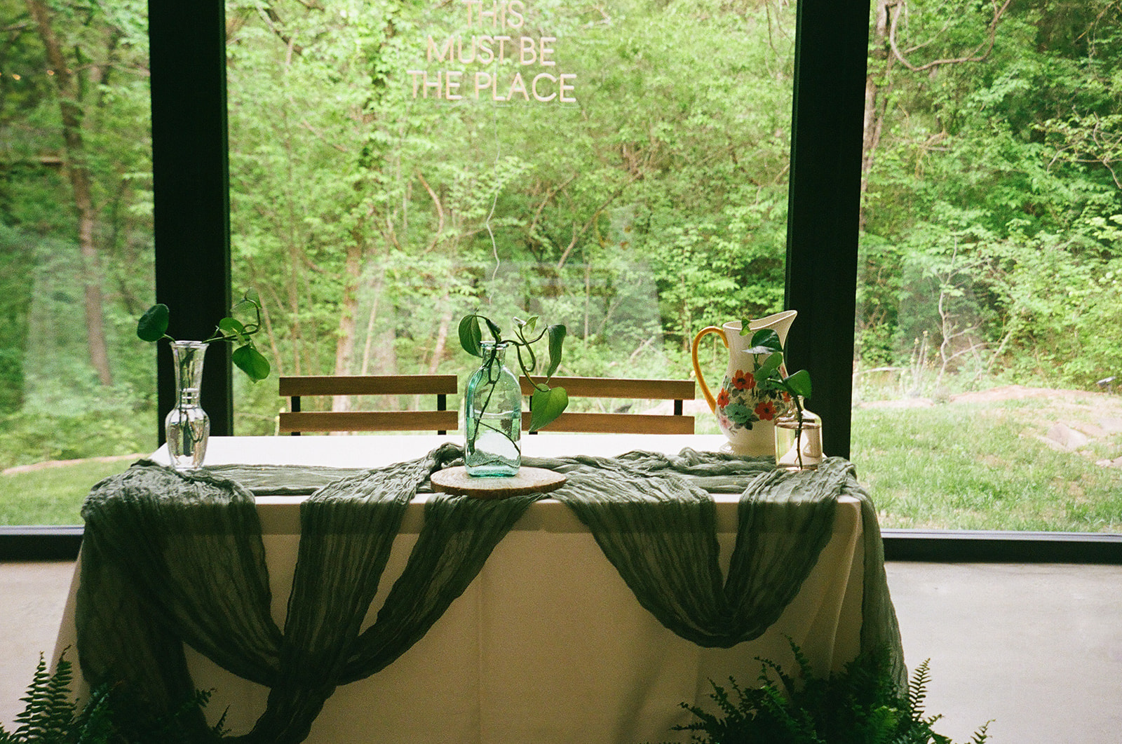 Rectangular tables with white tablecloths and green runners are arranged in a bright room with large windows and wooden chairs, overlooking a green outdoor view.