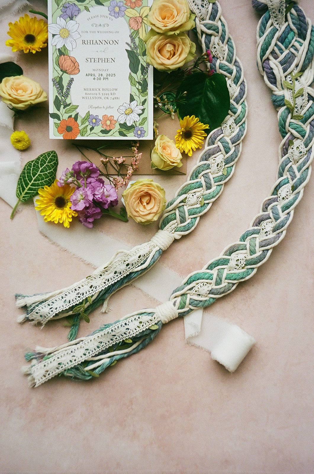 Two floral wedding invitations surrounded by yellow and purple flowers, green leaves, a wax seal, and a decorative box arranged on a light pink surface.