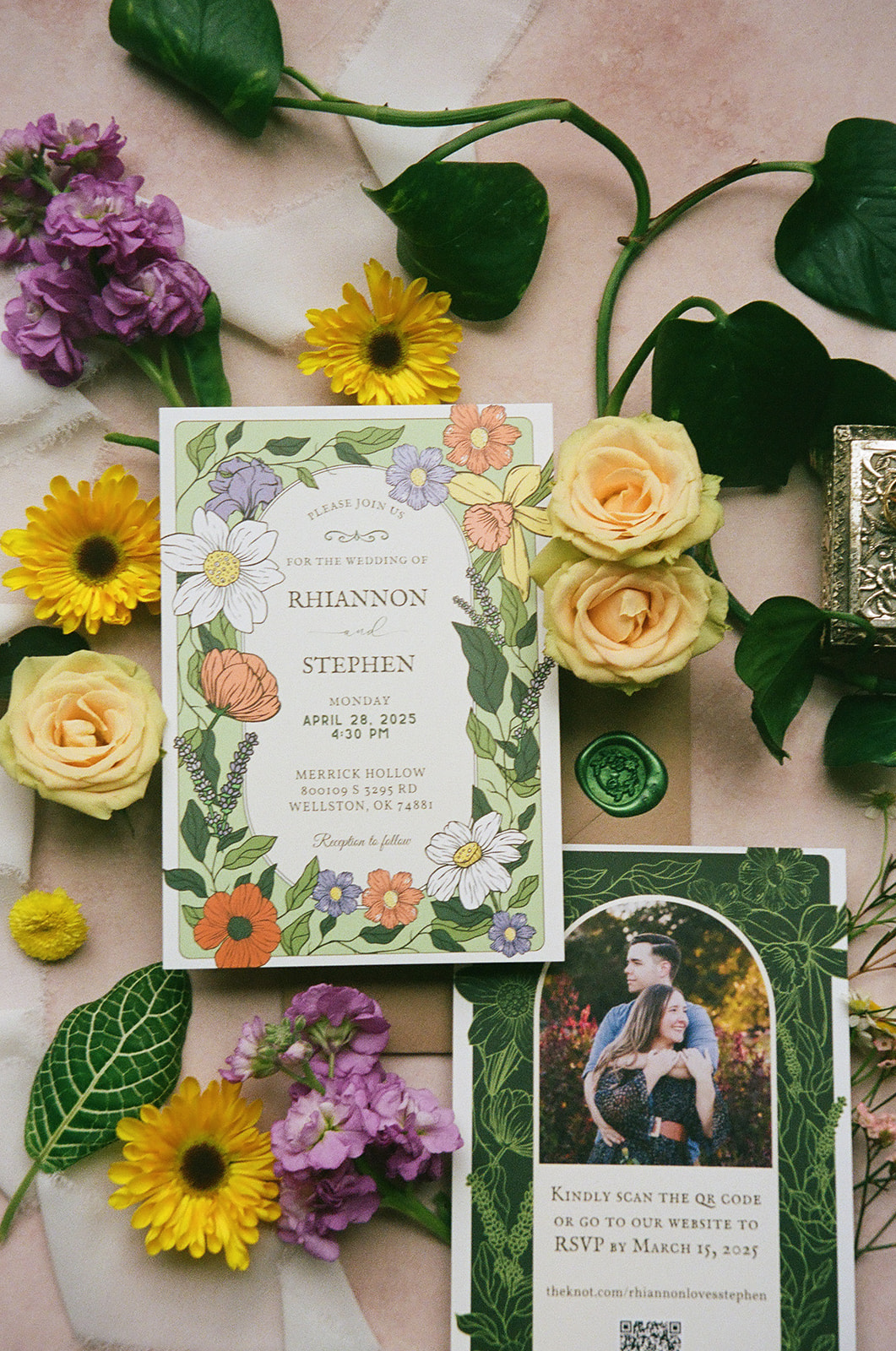 Two floral wedding invitations surrounded by yellow and purple flowers, green leaves, a wax seal, and a decorative box arranged on a light pink surface.