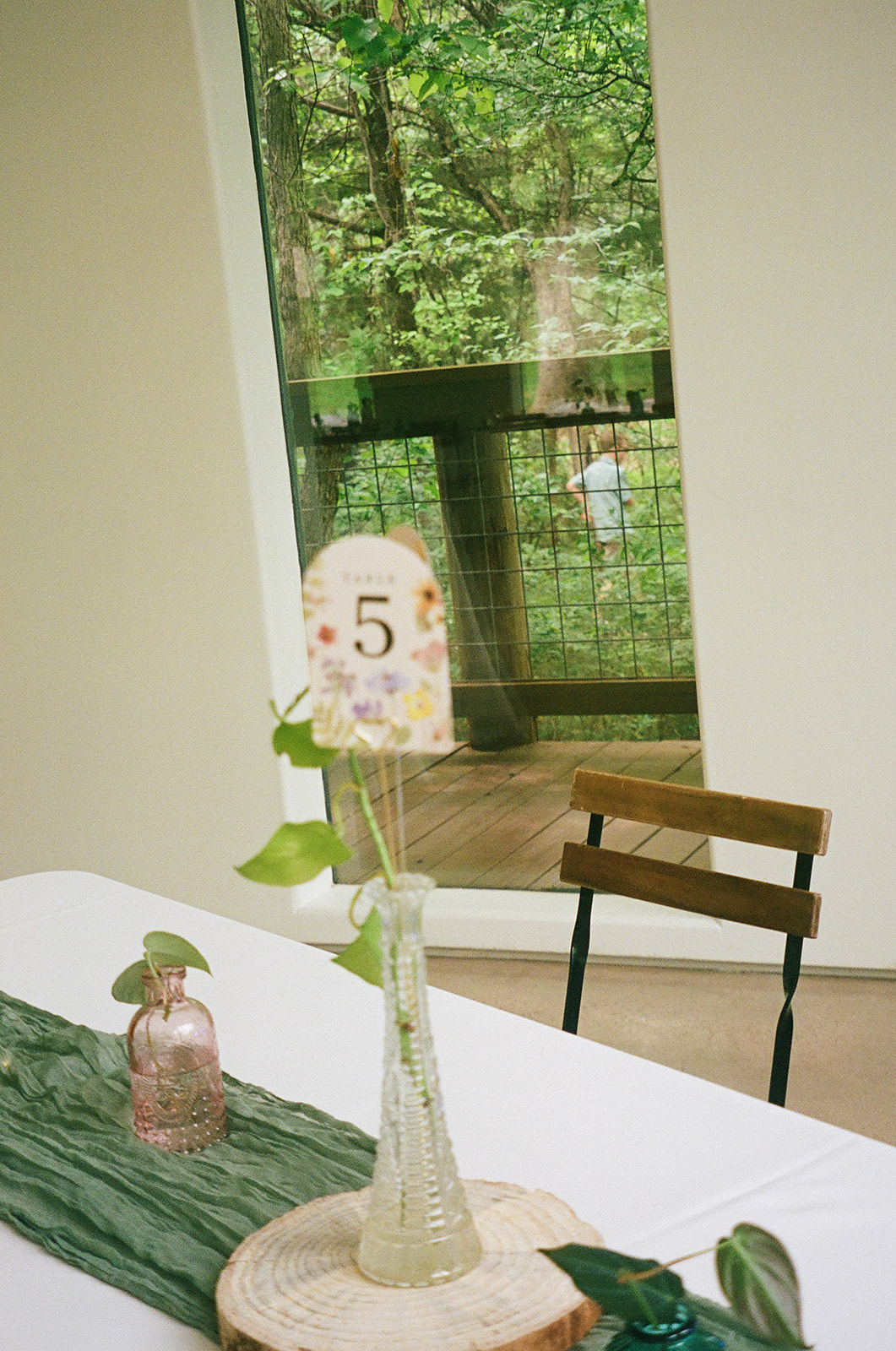 Rectangular tables with white tablecloths and green runners are arranged in a bright room with large windows and wooden chairs, overlooking a green outdoor view.