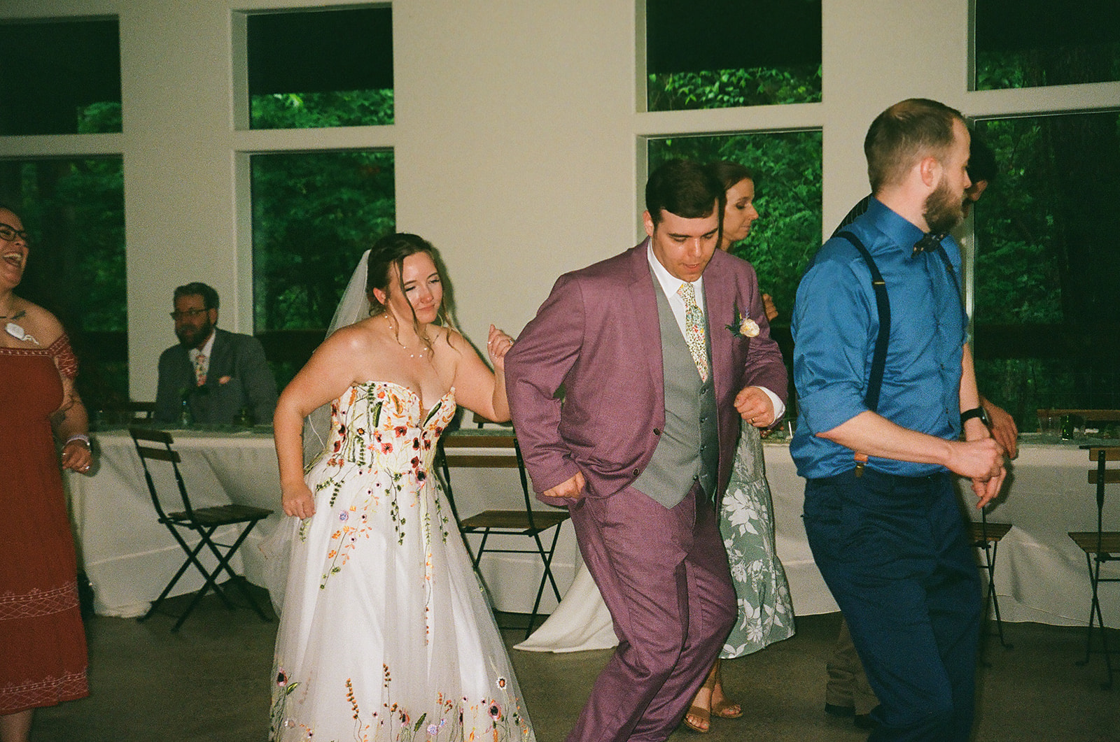 A bride in a white dress and a groom in a lavender suit dance with guests indoors at a daytime wedding reception.