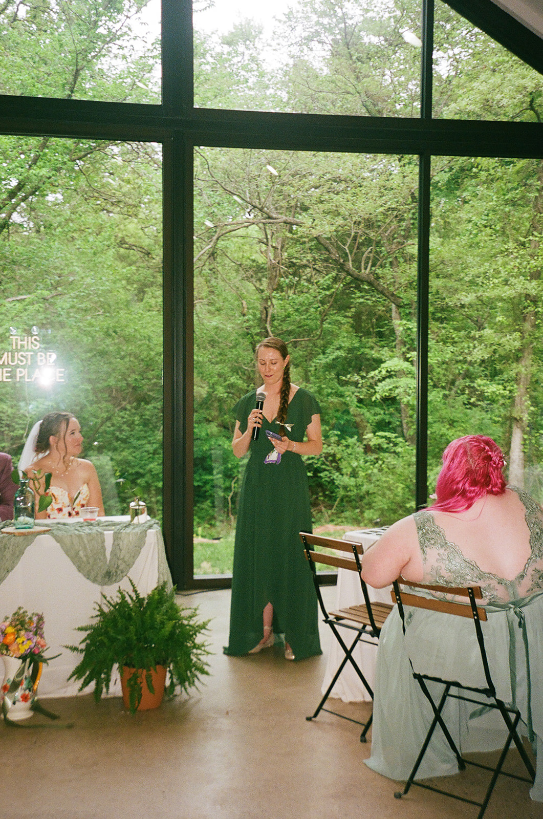 A woman in a green dress stands and speaks into a microphone at a wedding reception; two other women sit and listen near large windows with a forest view for their wedding at Merrick Hollow 