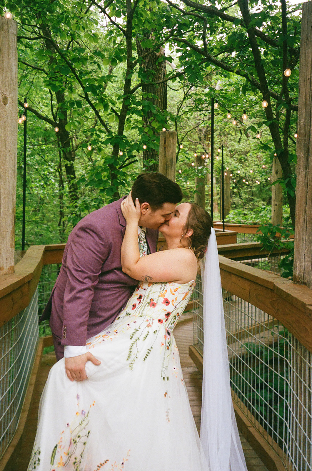 A groom in a purple suit kisses the bride’s forehead as they embrace outdoors, surrounded by greenery. The bride is draped in a white veil and holds a colorful bouquet for their wedding at Merrick Hollow 