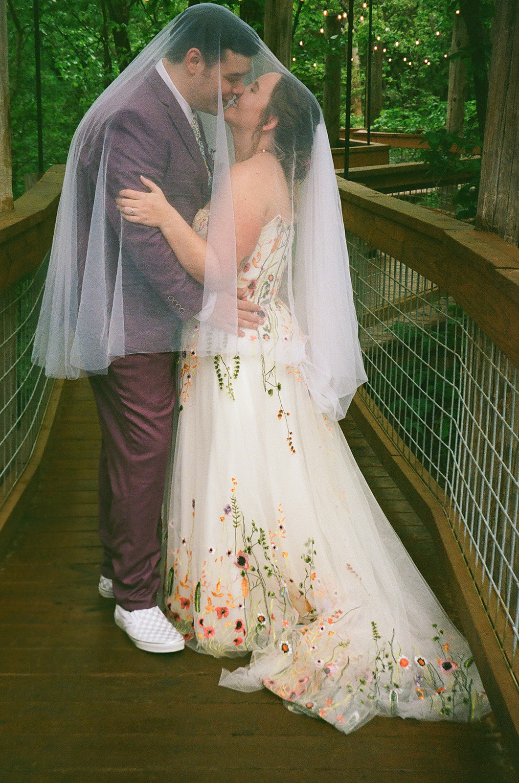 A groom in a purple suit kisses the bride’s forehead as they embrace outdoors, surrounded by greenery. The bride is draped in a white veil and holds a colorful bouquet for their wedding at Merrick Hollow 