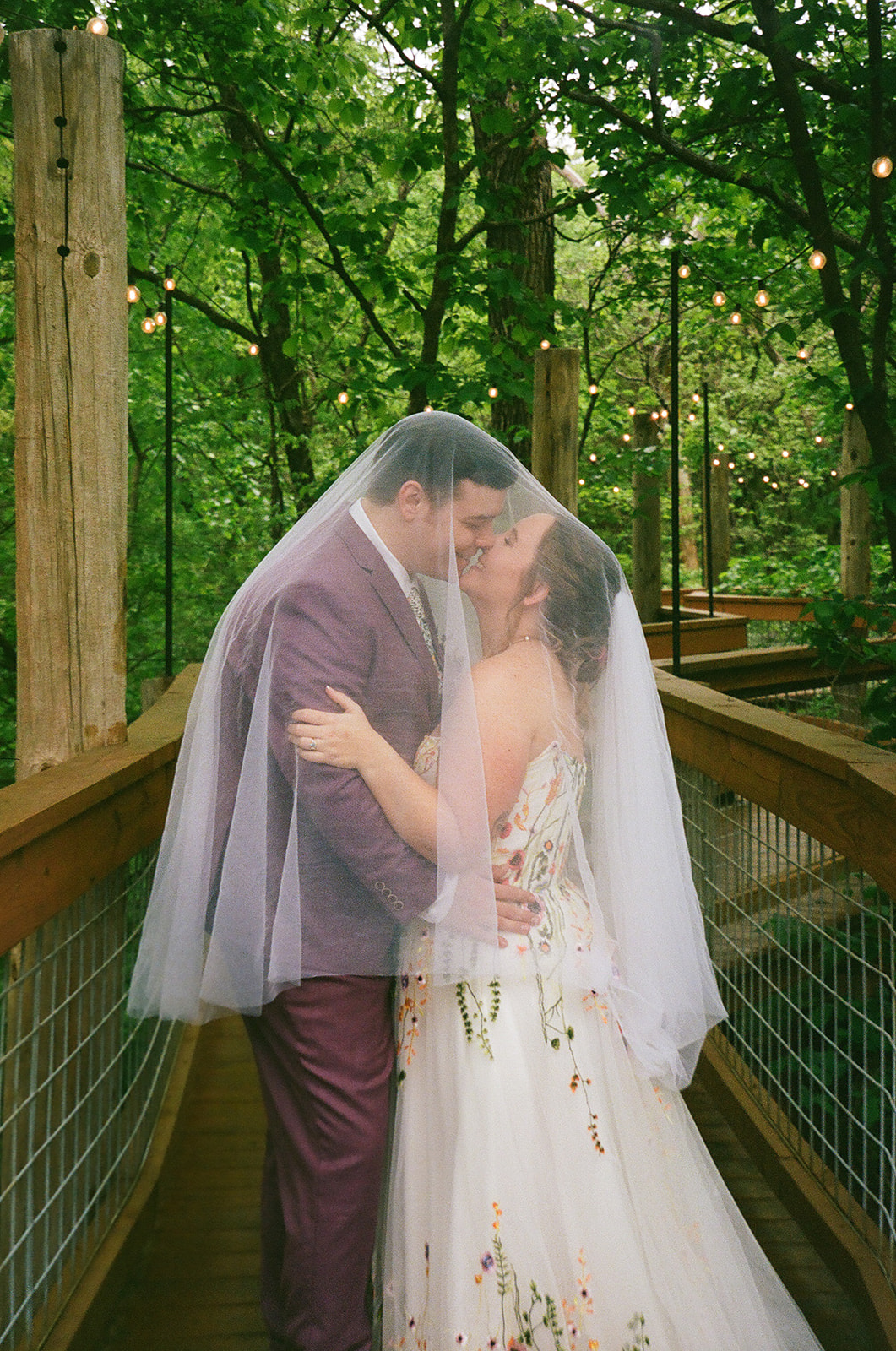 A groom in a purple suit kisses the bride’s forehead as they embrace outdoors, surrounded by greenery. The bride is draped in a white veil and holds a colorful bouquet for their wedding at Merrick Hollow 