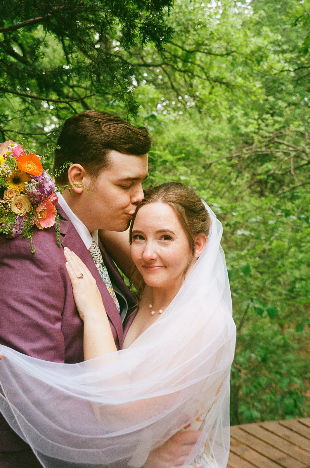 A groom in a purple suit kisses the bride’s forehead as they embrace outdoors, surrounded by greenery. The bride is draped in a white veil and holds a colorful bouquet for their wedding at Merrick Hollow 