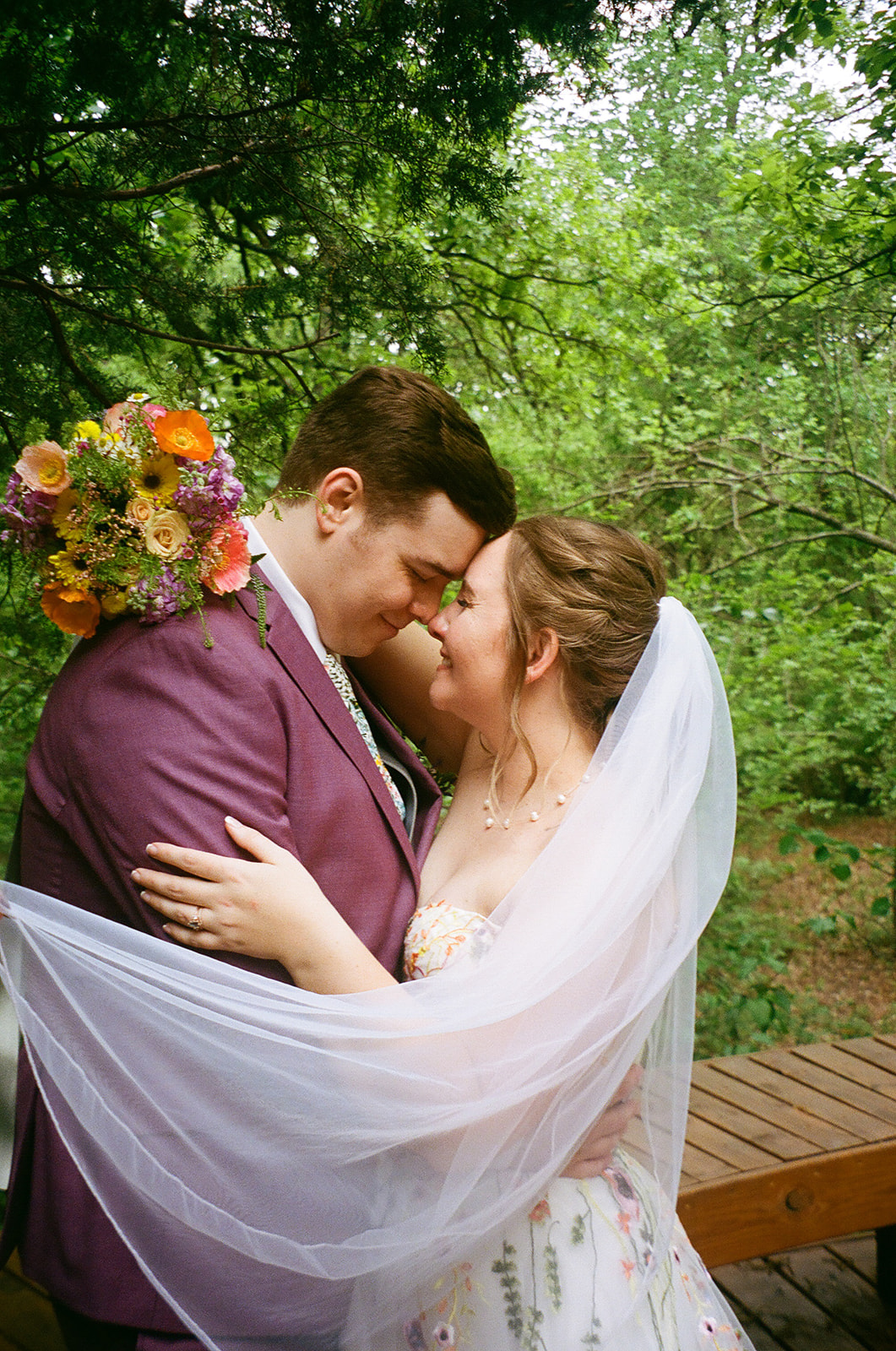 A groom in a purple suit kisses the bride’s forehead as they embrace outdoors, surrounded by greenery. The bride is draped in a white veil and holds a colorful bouquet for their wedding at Merrick Hollow 