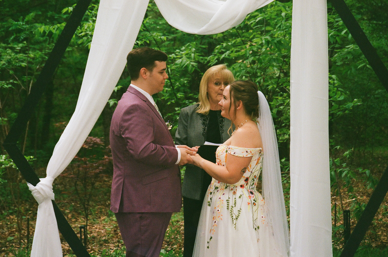 A bride and groom stand facing each other, holding hands during an outdoor wedding ceremony, with an officiant and greenery in the background for their wedding at Merrick Hollow 