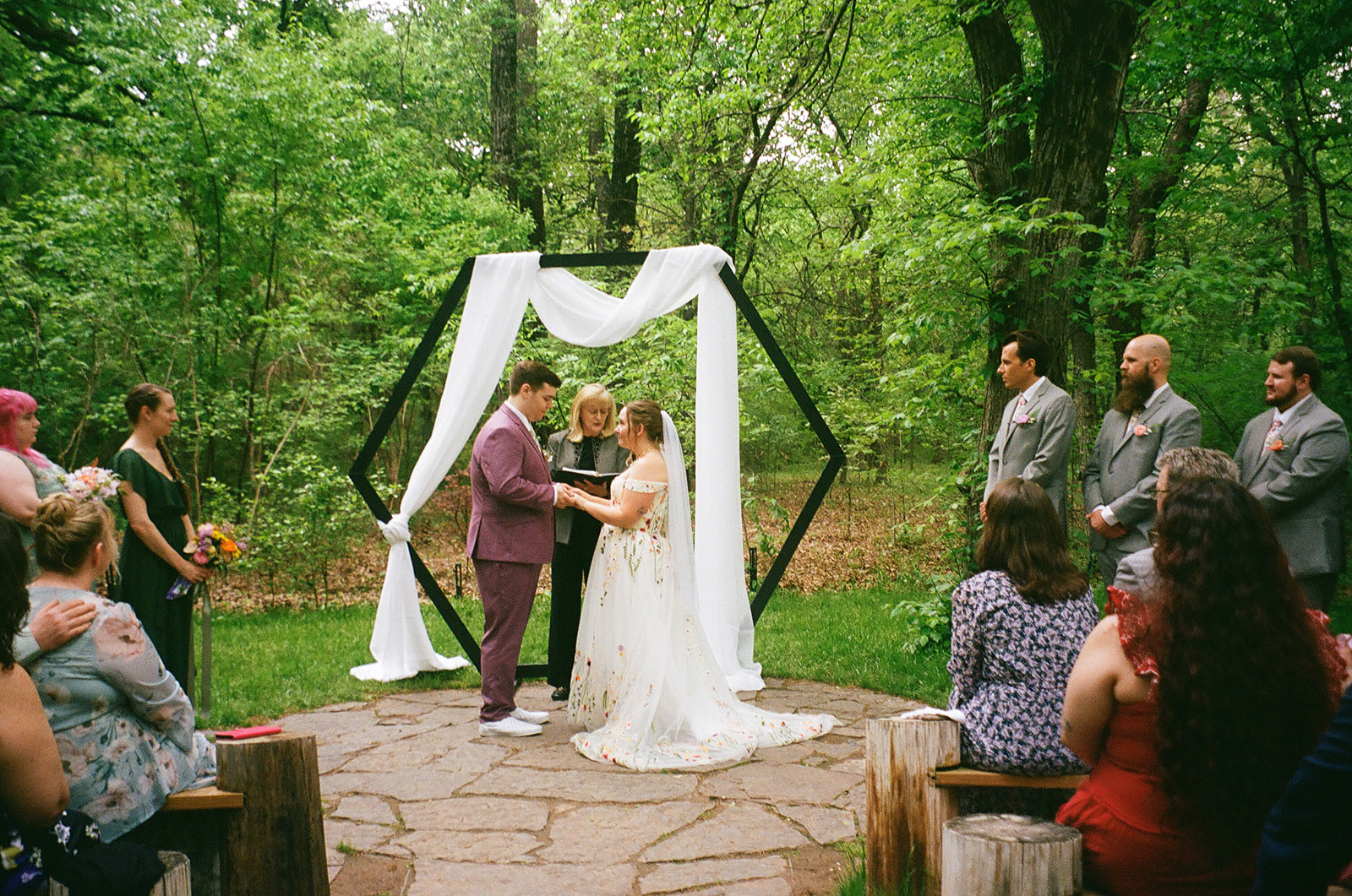 A bride and groom stand facing each other, holding hands during an outdoor wedding ceremony, with an officiant and greenery in the background for their wedding at Merrick Hollow 
