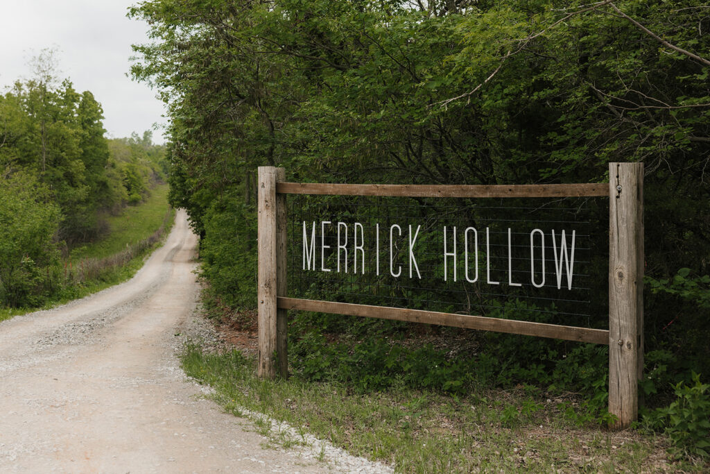 A wooden sign reading "Merrick Hollow" stands beside a gravel road surrounded by green trees and grass