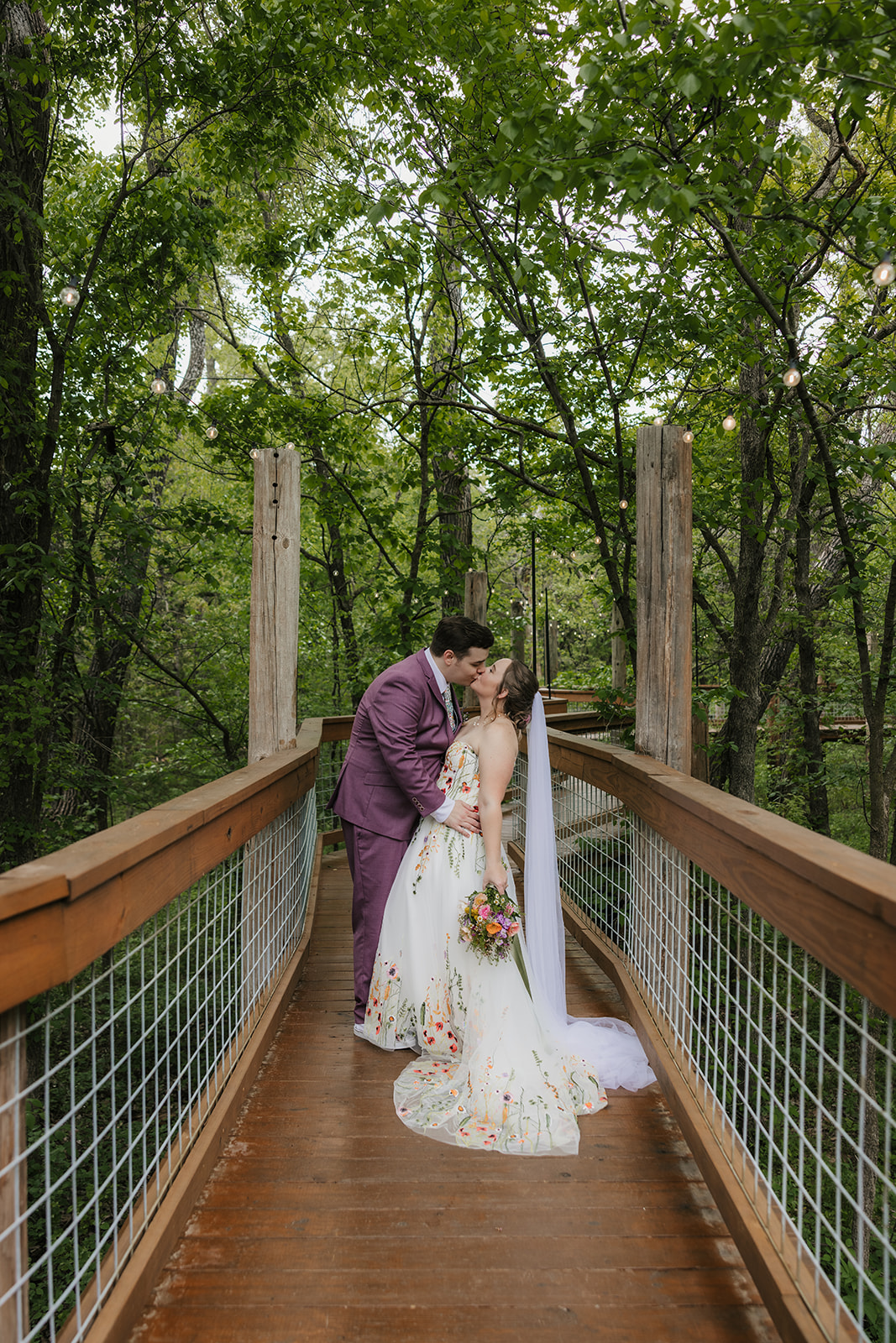 A bride and groom embrace on a wooden deck surrounded by greenery; the groom wears a mauve suit and the bride wears a white dress with floral details and a veil.