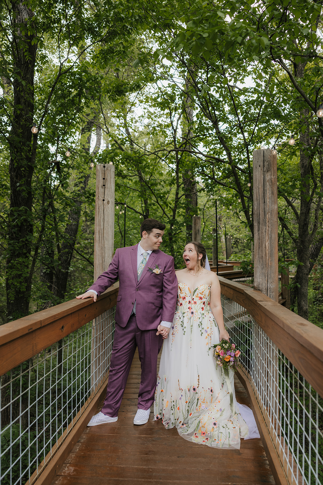 A bride and groom embrace on a wooden deck surrounded by greenery; the groom wears a mauve suit and the bride wears a white dress with floral details and a veil.