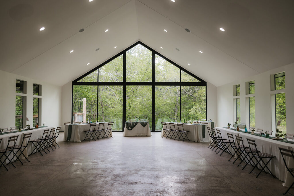 A spacious, modern event hall with large triangular windows, folding chairs, and decorated tables lined along both sides, overlooking green trees outside.