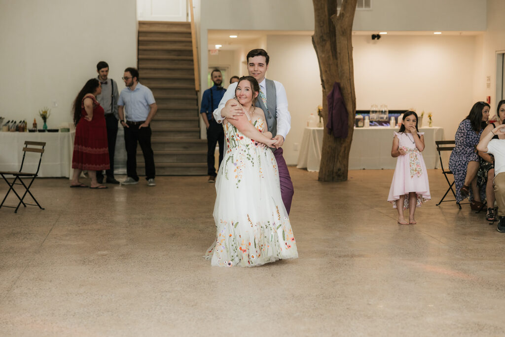 A young girl in a pink dress spins barefoot on a polished floor while several adults and another child are in the background at an indoor gathering.