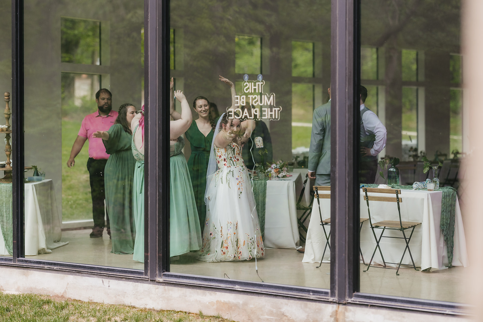 A group of people, including a bride in a white dress, are gathered inside a glass-walled building, some with raised arms, seen through large windows.